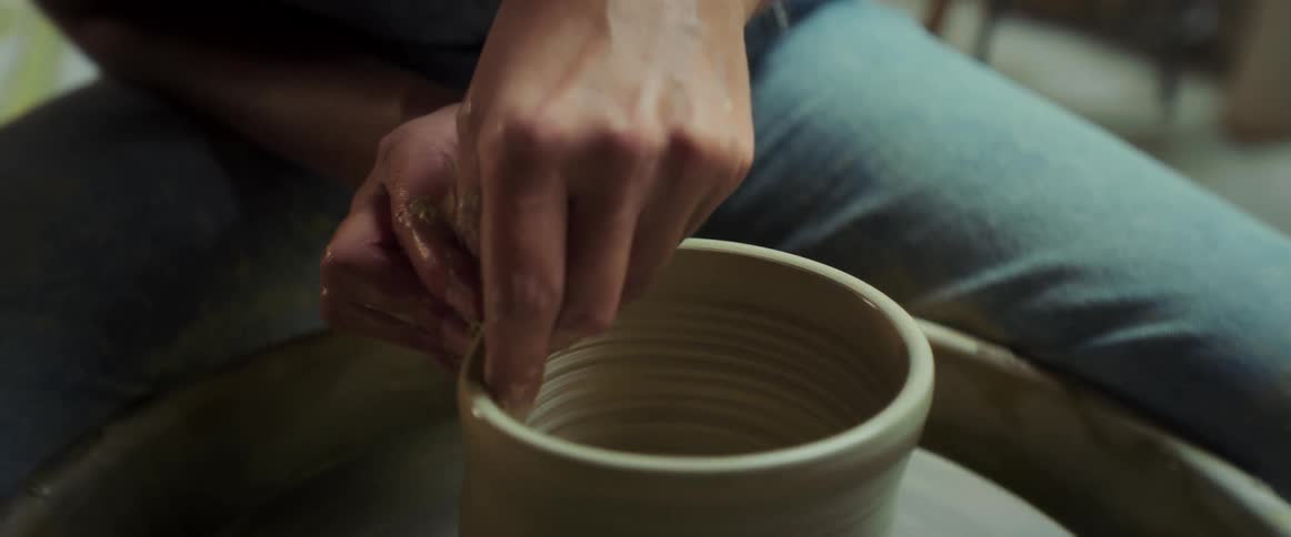 Potter sitting at pottery wheel shapes clay vessel
