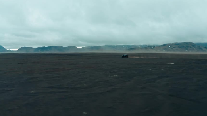 A lone vehicle drives through a vast dark volcanic sand plain with distant mountains