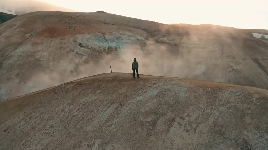 A person stands on a barren hill with steam rising from the ground