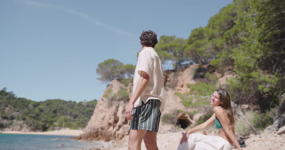 A man stands on a beach next to a woman sitting on a rock
