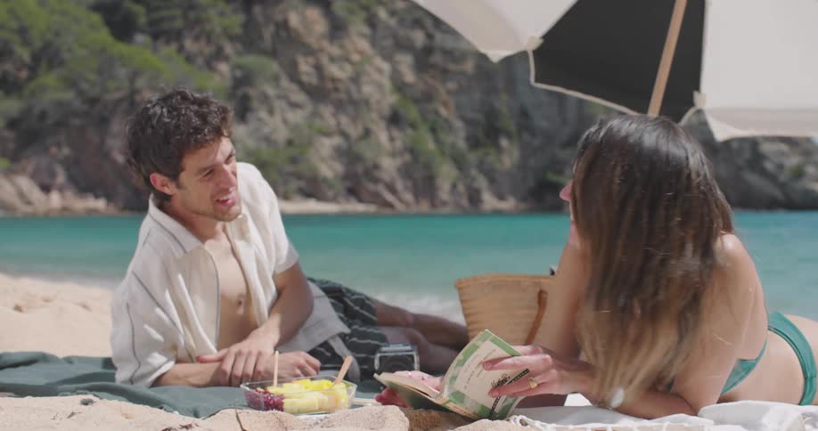 A young couple lying on a sandy beach under a black and white umbrella