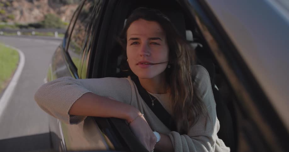 A woman with long hair in the passenger seat of a car arm resting on the window sill