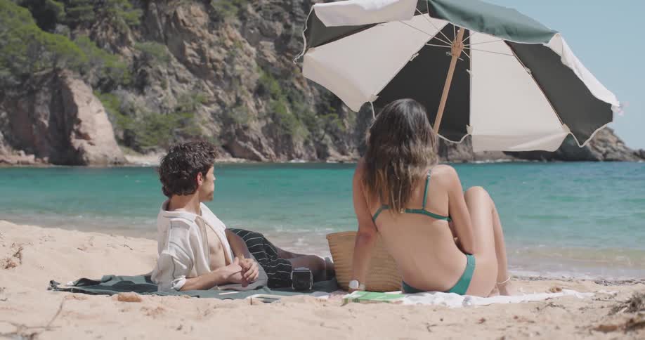 Woman offering a toothpick with fruit to her husband while they are sitting on the sand on a turquoise beach
