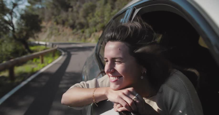 A woman smiling with excitement waves happily from the front seat of a car on a coastal road