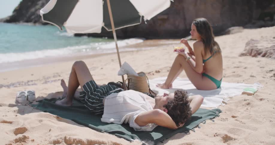 Man lying on towel on the beach sand next to his wife who is eating fruit