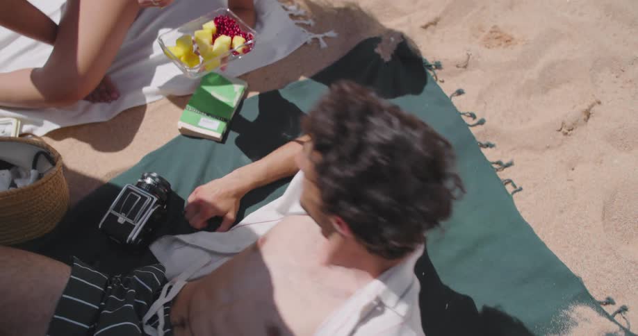 A young man resting on the beach eats a piece of fruit offered to him