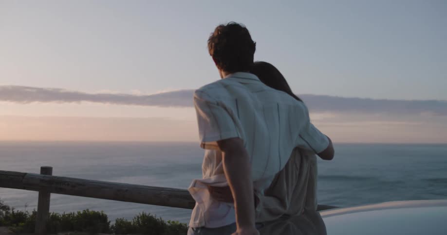 A young couple seated on the front of a car facing the ocean at sunset