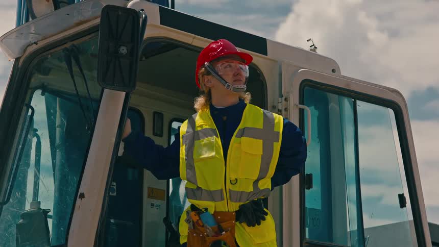 Woman in a yellow highvisibility vest and red hard hat gets out of a construction vehicle cabin