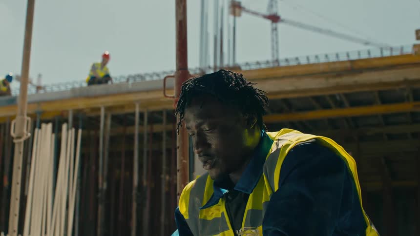 A construction worker in a yellow highvisibility jacket and blue shirt is sweeping sweat from his face with his arm