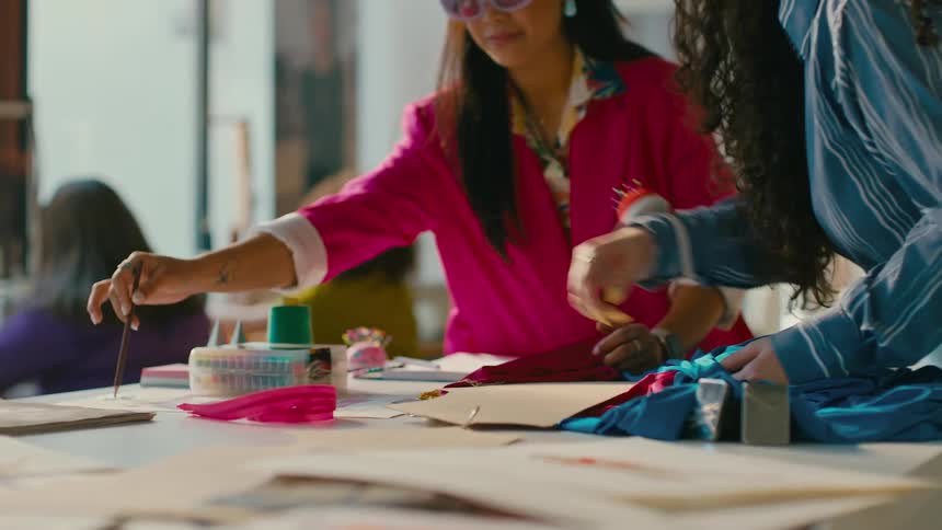 An fashion designer wearing a red hat and pink jacket stands at a table with fabrics