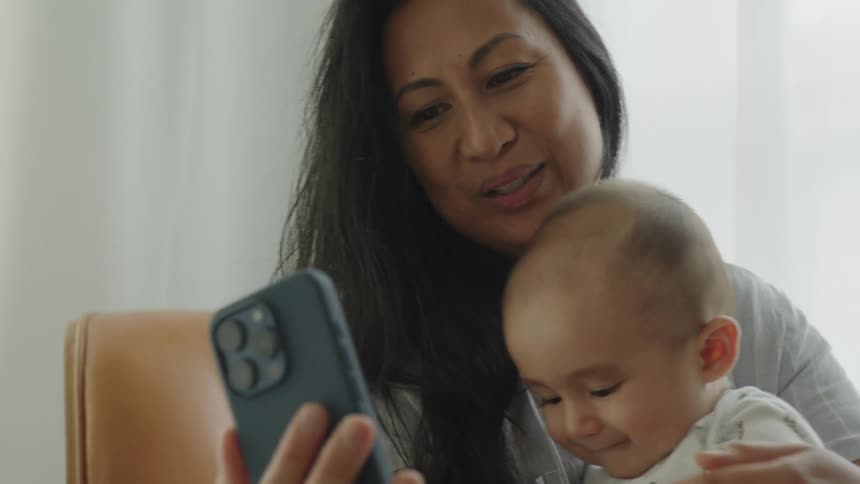 A mother holding her baby while looking at a smartphone making a video call