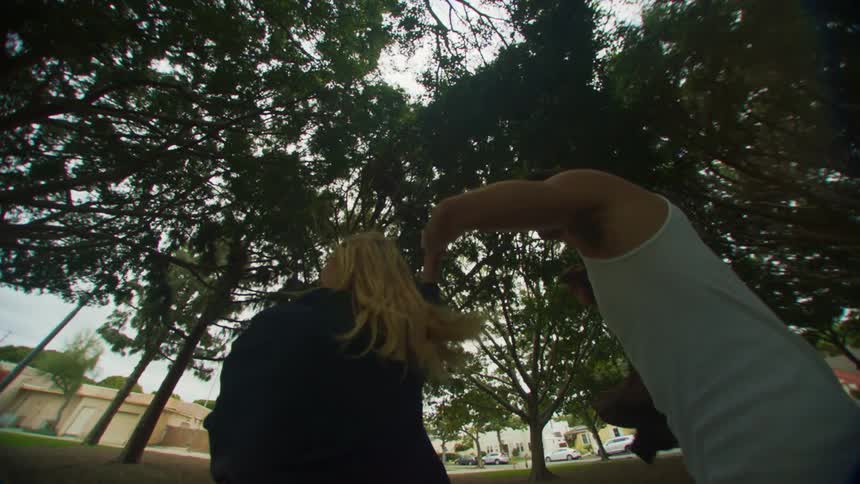 A blonde woman wearing a black jacket plays and dances on the green lawn in the public park