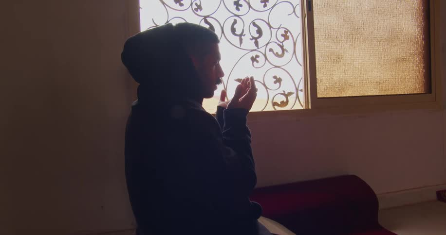 A young man pushes his hood back and begins praying in a mosque