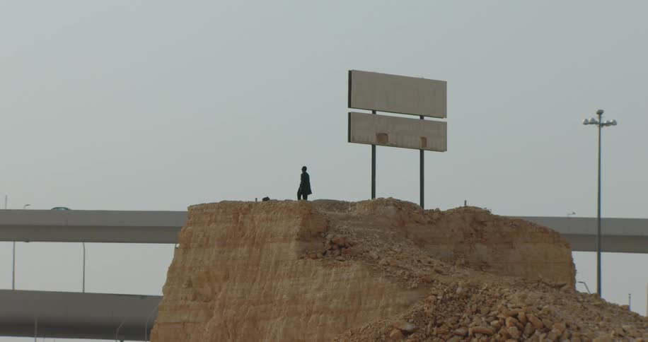 A person stands on a mound of earth beside an empty billboard close to a highway