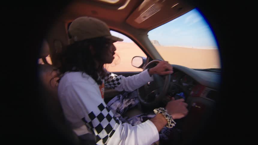 Man with wavy hair a cap and a white shirt drives a car in the desert