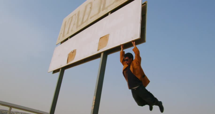 A young man in an orange jacket jumps up and grabs a billboard near a highway hanging from it for a few seconds before letting go
