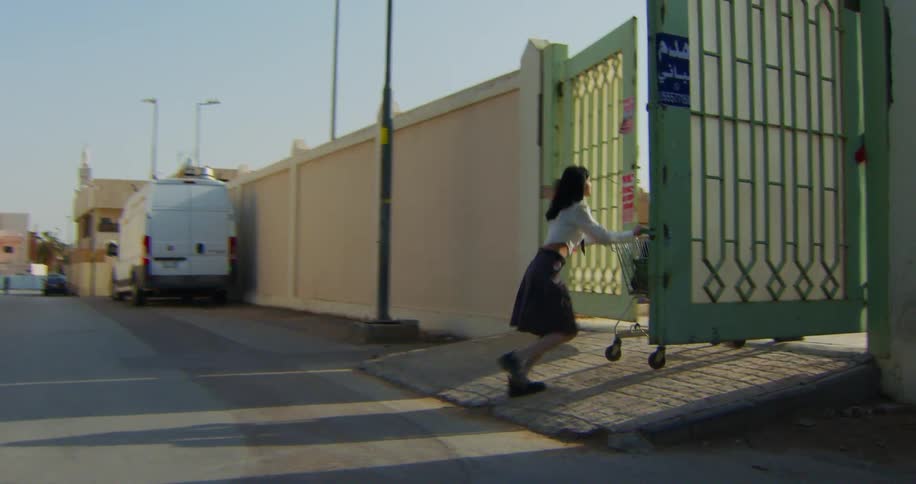 An asian woman rolls a grocery cart up a ramp and through a gate