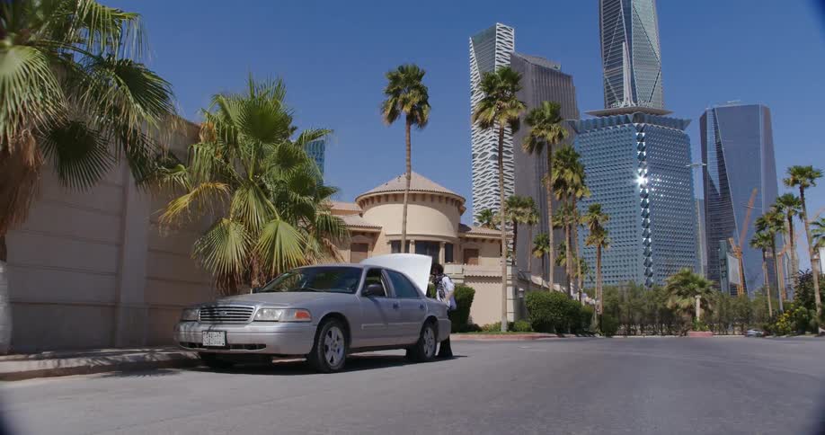 Two people in the trunk of a silver car parked on a street in a metropolis with a modern skyscraper in the background