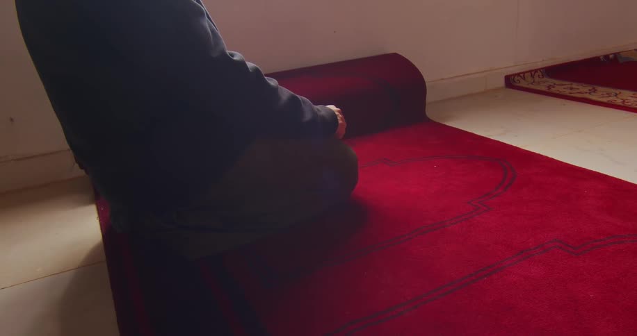 A person is facing backwards wearing a black hooded coat and is kneeling on a red carpet