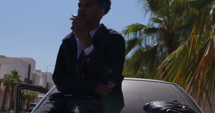 A black man sits on a car while smoking a cigarette