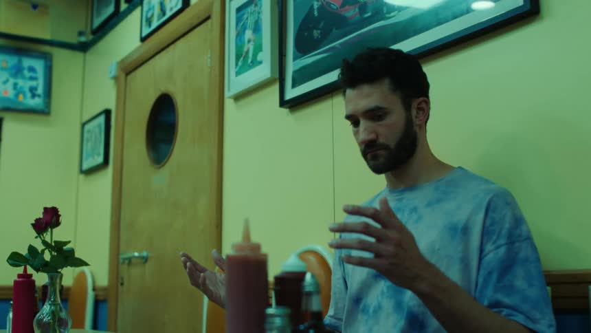 Bearded man wearing a blue t shirt looks down on his hands in a room
