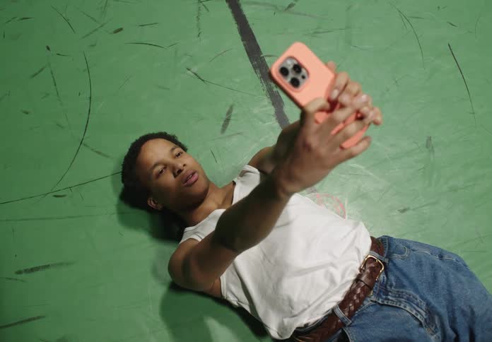 A man wearing a white undershirt a belt and jeans lies down on the green ground taking a selfie with a smartphone in the basketball court
