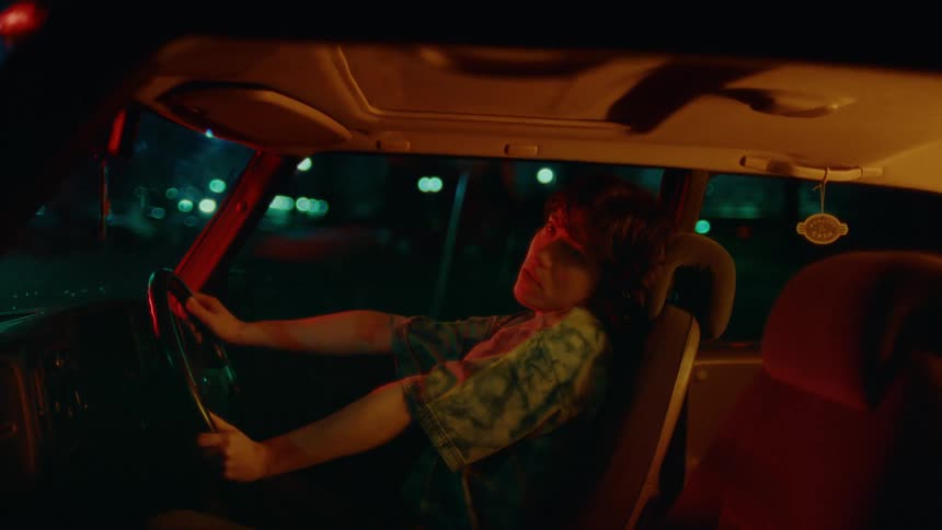 In the dim glow of the parking lot lights a woman wearing a blue tshirt is gripped by a seizure as she sits in her car