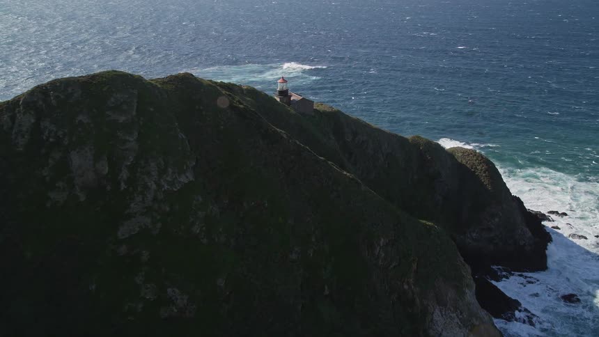 A white lighthouse with a red roof stands on a green cliff overlooking a vast blue ocean