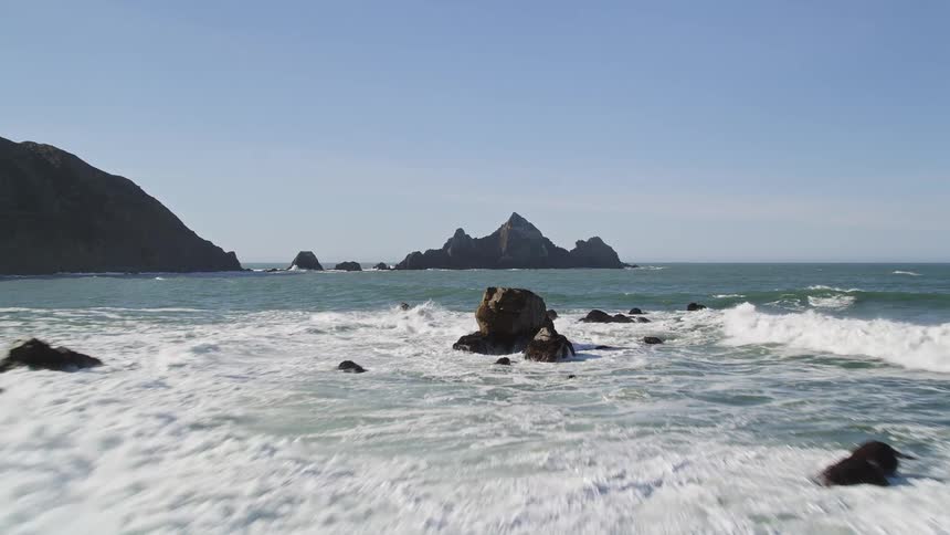 The ocean crashes against the rocky shore on a sunny day sending plumes of mist skyward