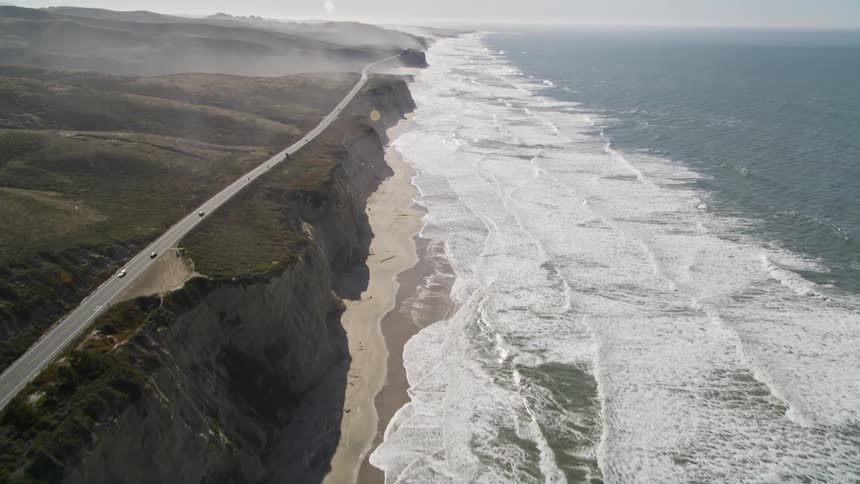 Aerial view of big ocean waves crashing against beach near the pacific coast highway running parallel to the vast ocean