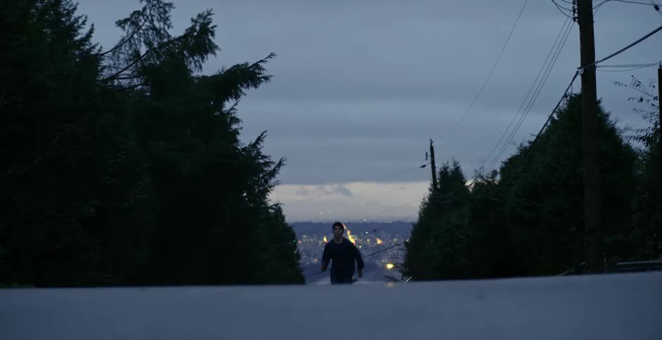 A young man running up a steep road with flames visible in the valley community behind him