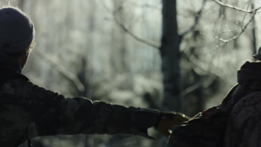 Clad in a wool hat and jacket arrows fletched across his back one hunter nudges his friend pointing towards an animal in the woods