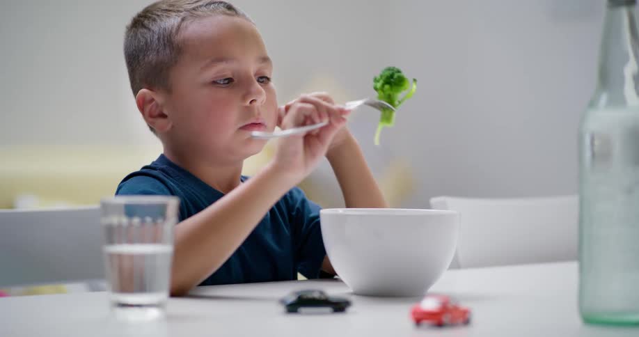 A child grimaces as he uses a fork to pick up some broccoli from a white bowl