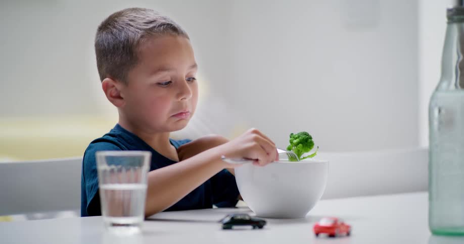 A little boy sitting at a table grimacing as he looks at some broccoli on the end of his fork