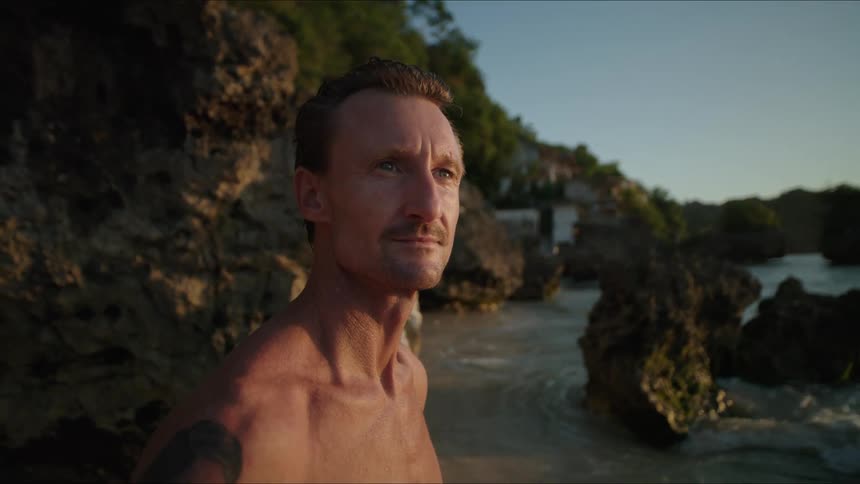 Man with a tattoo standing by big rocks looks around at the sea