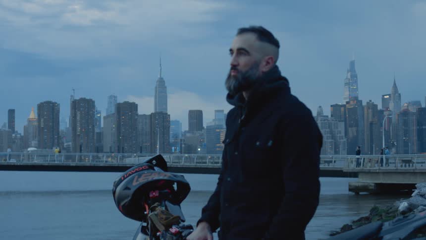 Man with motorcycle stands looking at the water while pedestrians walk along pier in the background