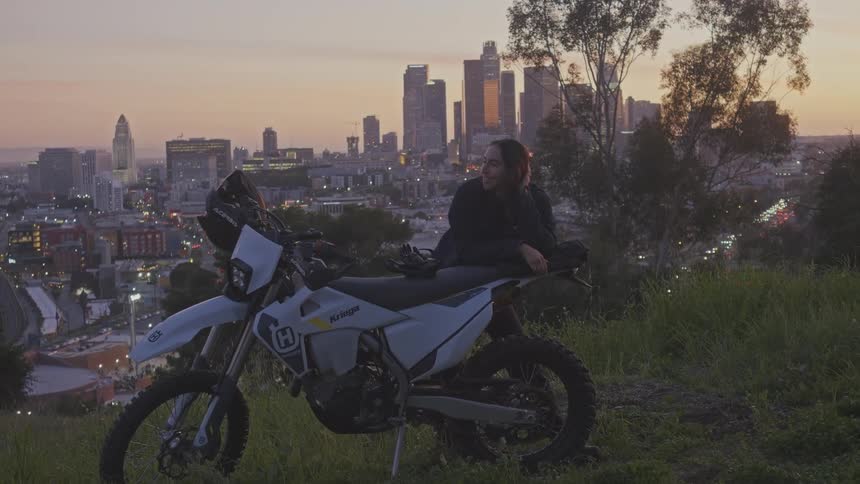 A woman leans on a white dirt bike at dusk overlooking a city skyline illuminated by the fading light of sunset