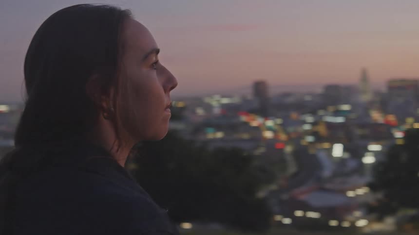 A young woman stands next to her motorcycle on a hillside overlooking the large city below