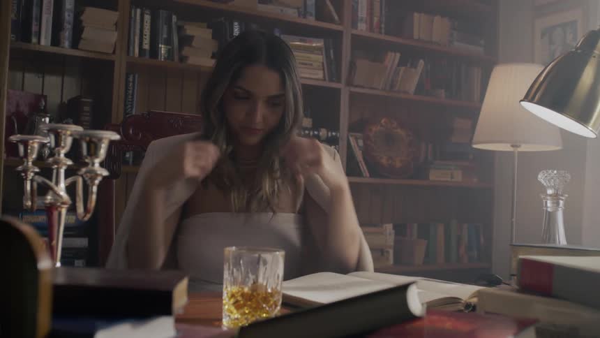 A long haired female is sitting at a desk flipping her hair and reading from a book