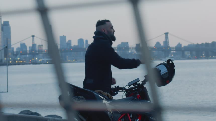 A man taking off his helmet and putting it on top of a motorcycle and then he puts the hood on his coat