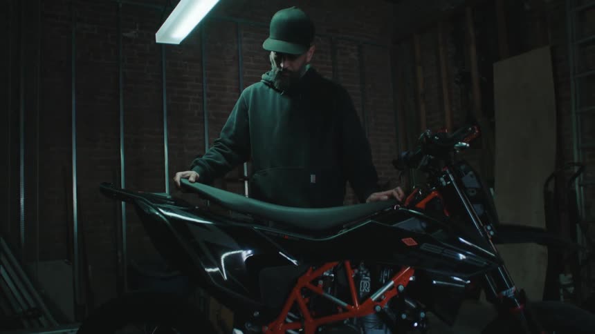 A man in a black coat and cap is putting a seat on a motorcycle