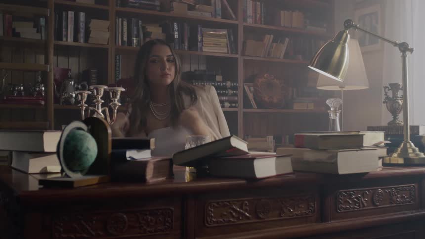 A young woman is sitting and in front of her there is a table with many books and she takes a glass of drink