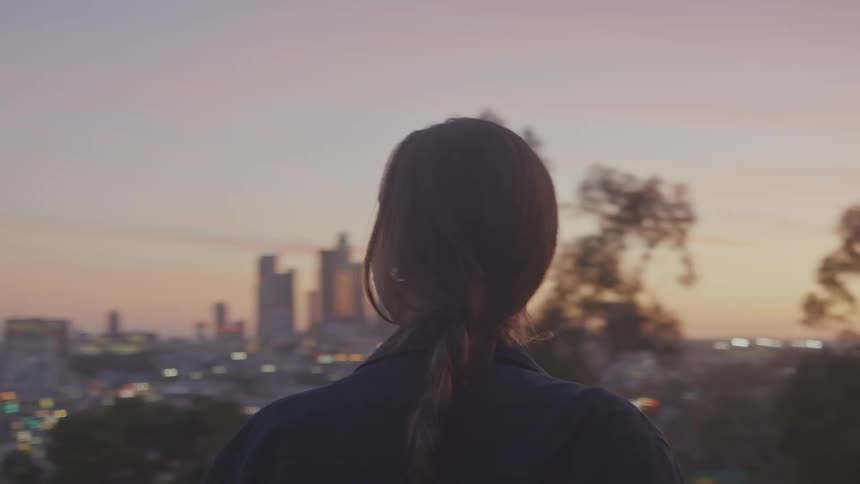 A woman reclined on a parked motorcycle looking at the city at sunset