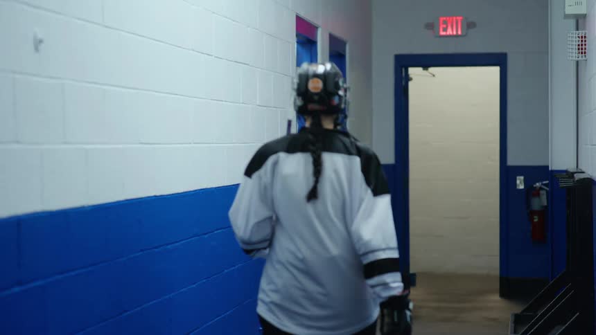 A person with hockey equipment is picking up a stick and walking down a corridor