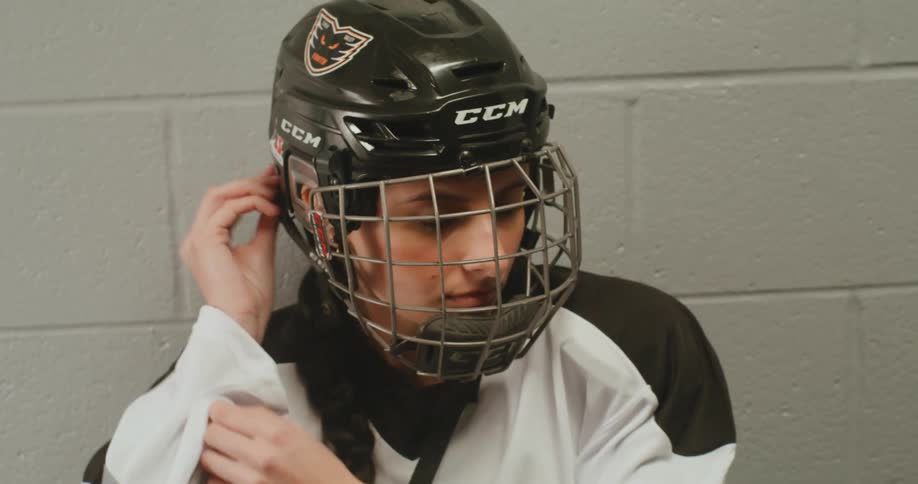 Female hockey player sitting against a white brick wall adjusting her helmet
