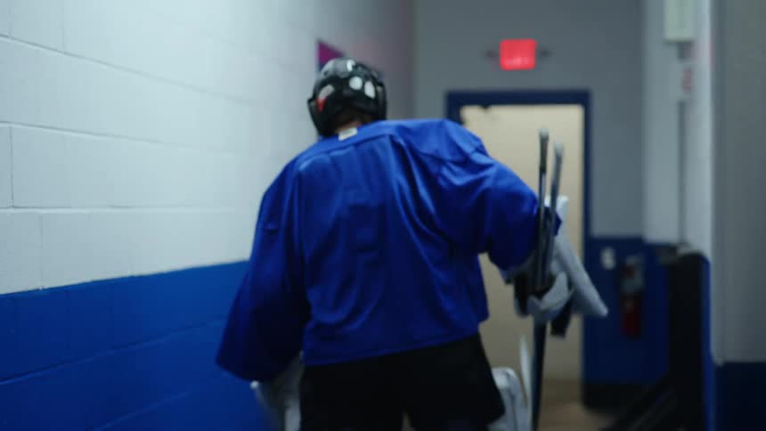 A hockey player grabs a stick and walks off down a hallway and through a door