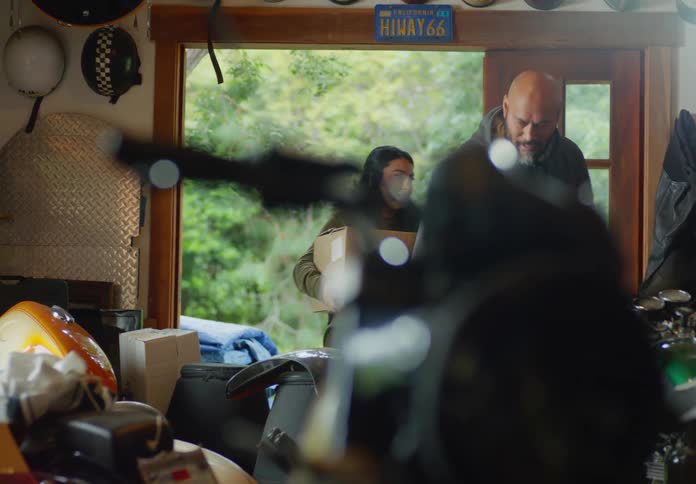 A bald, bearded man and a dark-haired woman are carrying a plastic bin and cardboard box into a messy storage shed.