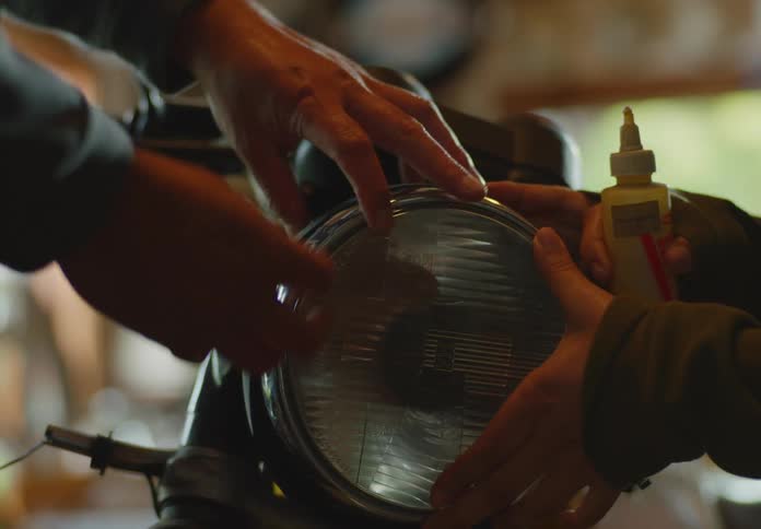 A dark-haired woman holds a bottle of glue and installs a new headlight on a motorcycle as the man offscreen helps hold it in place.