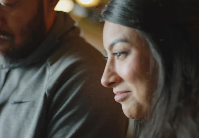 A dark skinned man with a bald head and beard and wearing a hoodie shows some tools and parts to a dark haired woman who squints and nods as they discuss.