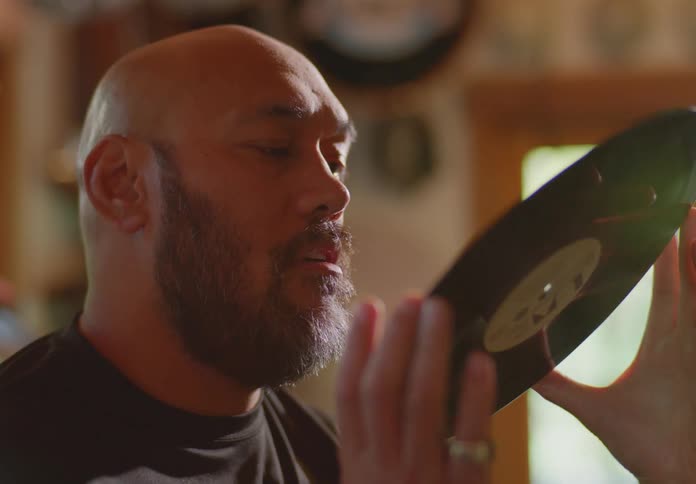 A bald and bearded ethnic man stands in a room with natural light and examines an old vinyl record.